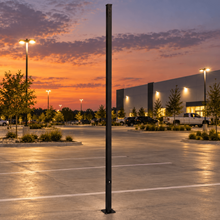 A 24' tall square straight black steel pole with a tenon mount stands vertically in the center of an empty parking lot at twilight, with  trees, parked cars, parking lot lights, and a building visible in the background under a colorful evening sunset sky.